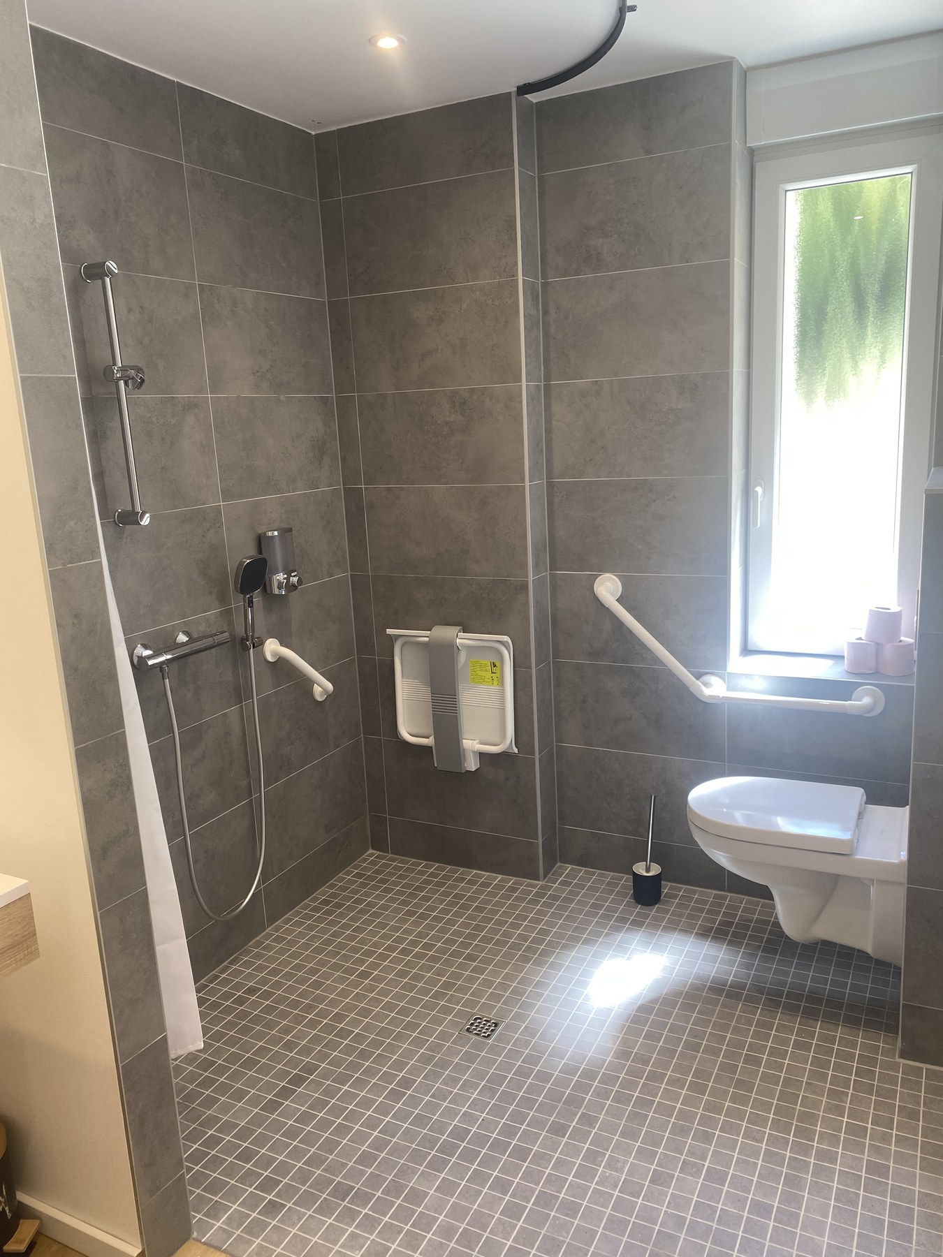Salle d'eau : douche à l'italienne avec siège mural relevable et barre d'appui, toilettes suspendues avec barre relevable, sol carrelé continu