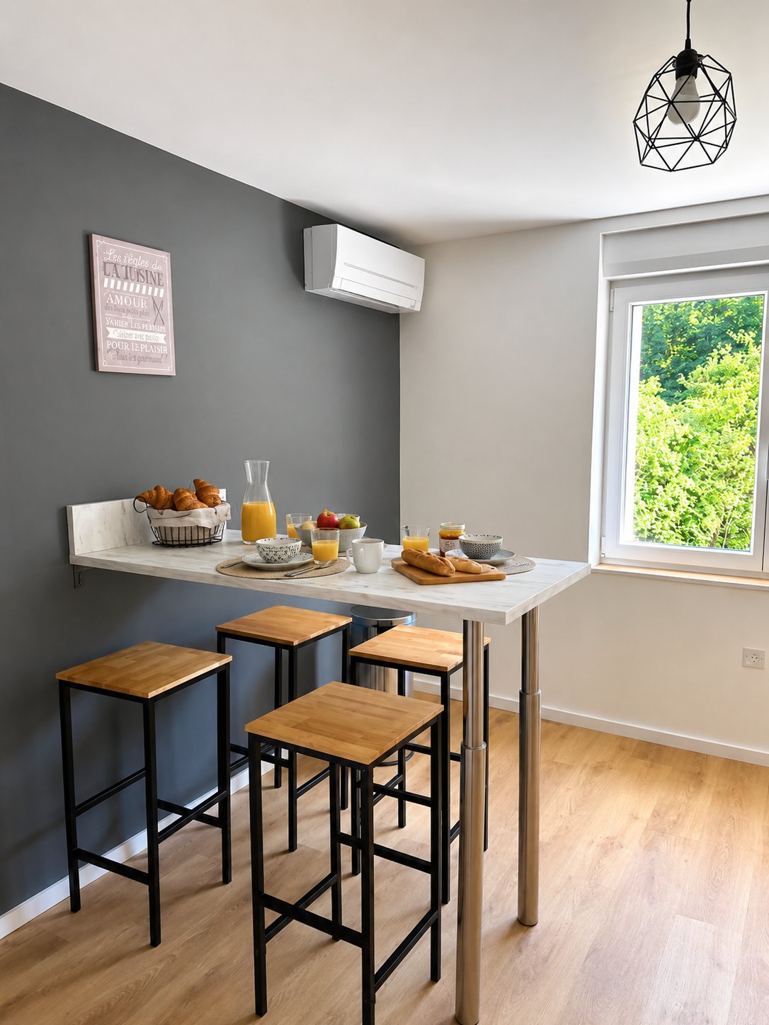 Table haute murale et quatre tabourets dans la cuisine, dressée pour le petit-déjeuner, climatiseur au mur et fenêtre sur la verdure